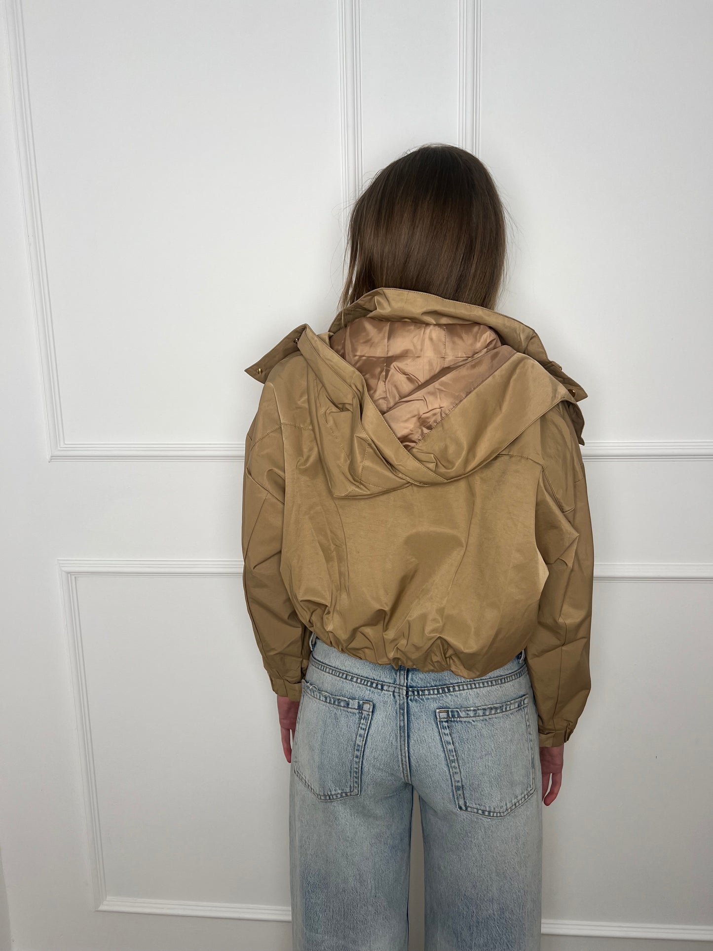 Frau steht vor weißer Wand und ist von hinten zu sehen und trägt eine beige cropped Jacke und eine Jeans