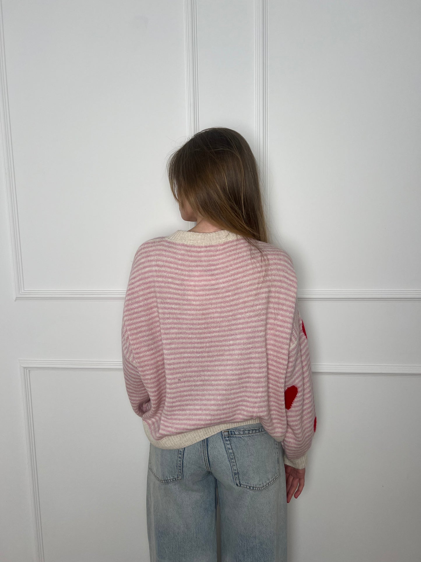 Frau vor weißer Wand von hinten trägt einen pink und weiss gestreiften Pullover mit roten Herzen.