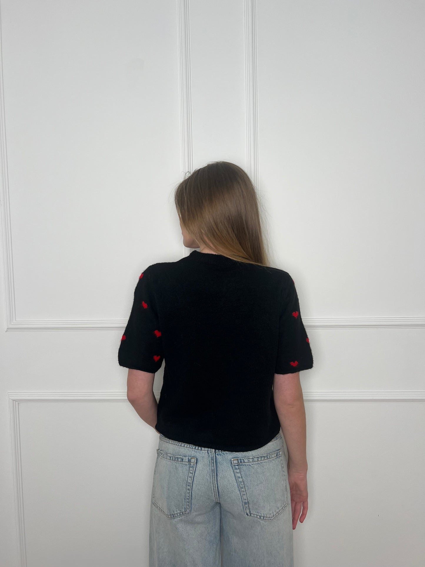 Frau von hinten vor weißer Wand trägt einen schwarzen Pullover mit kurzen Armen.