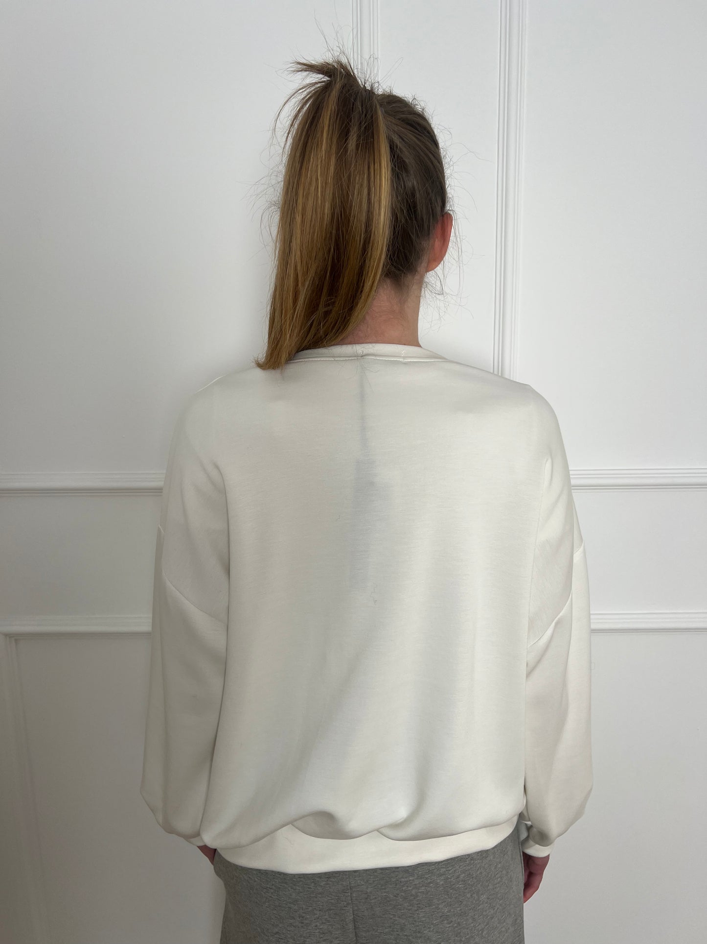 Frau vor weißer Wand trägt ein weißes Sweatshirt und ist von hinten zu sehen.