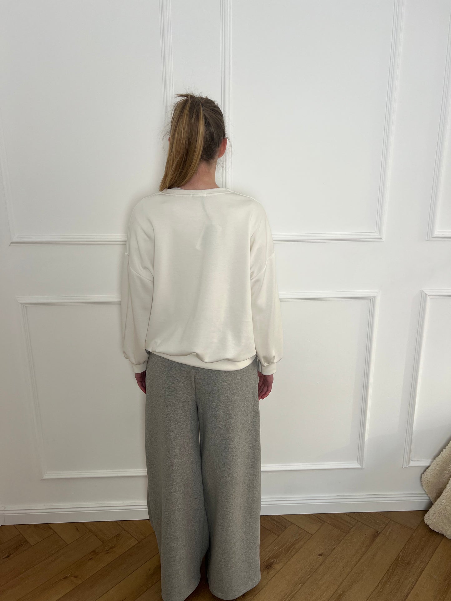 Frau vor weißer Wand trägt ein cremefarbenes Sweatshirt und eine graue Jogginghose