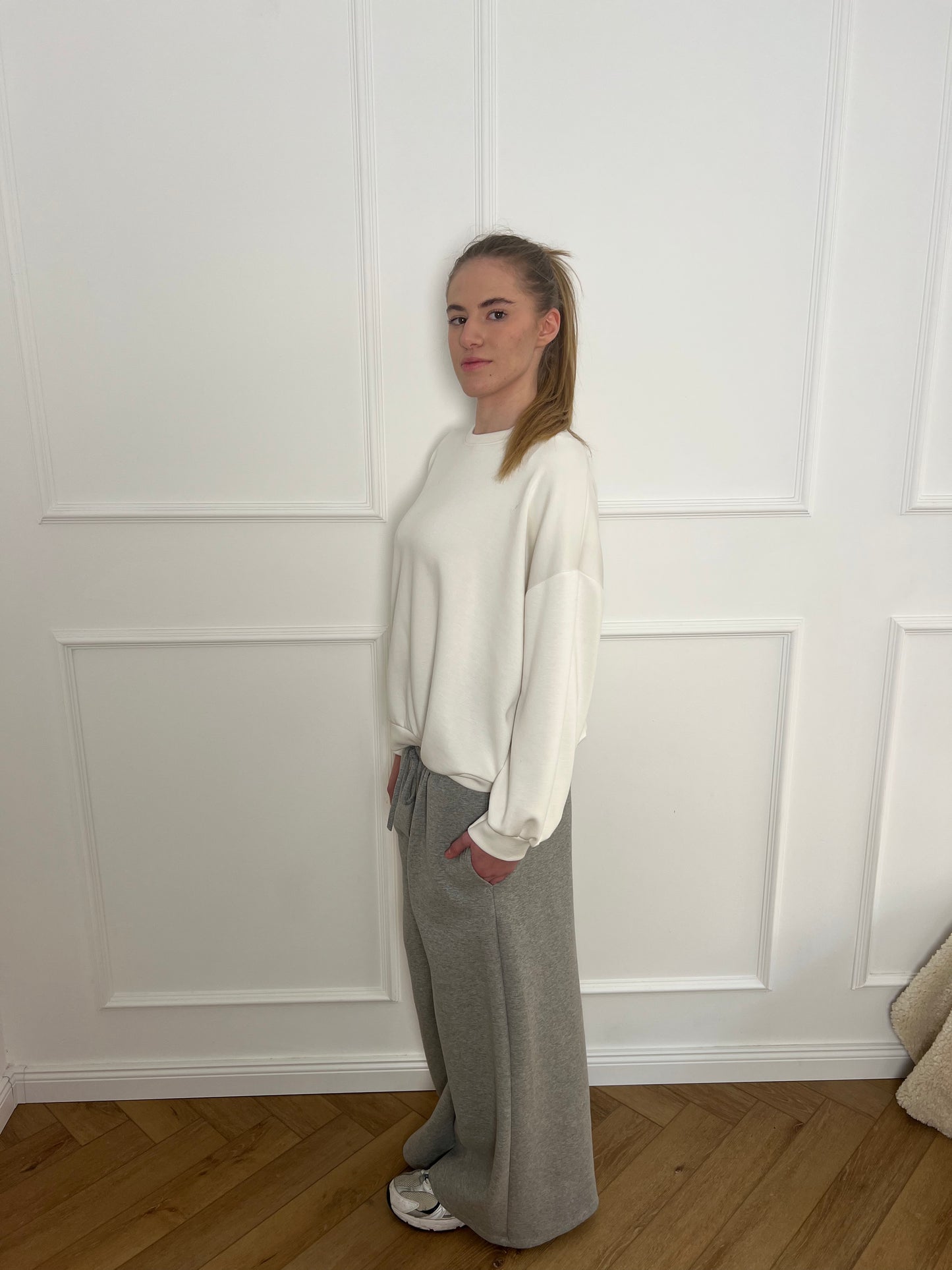 Frau vor weißer Wand trägt ein weißes Sweatshirt und eine graue Jogginghose