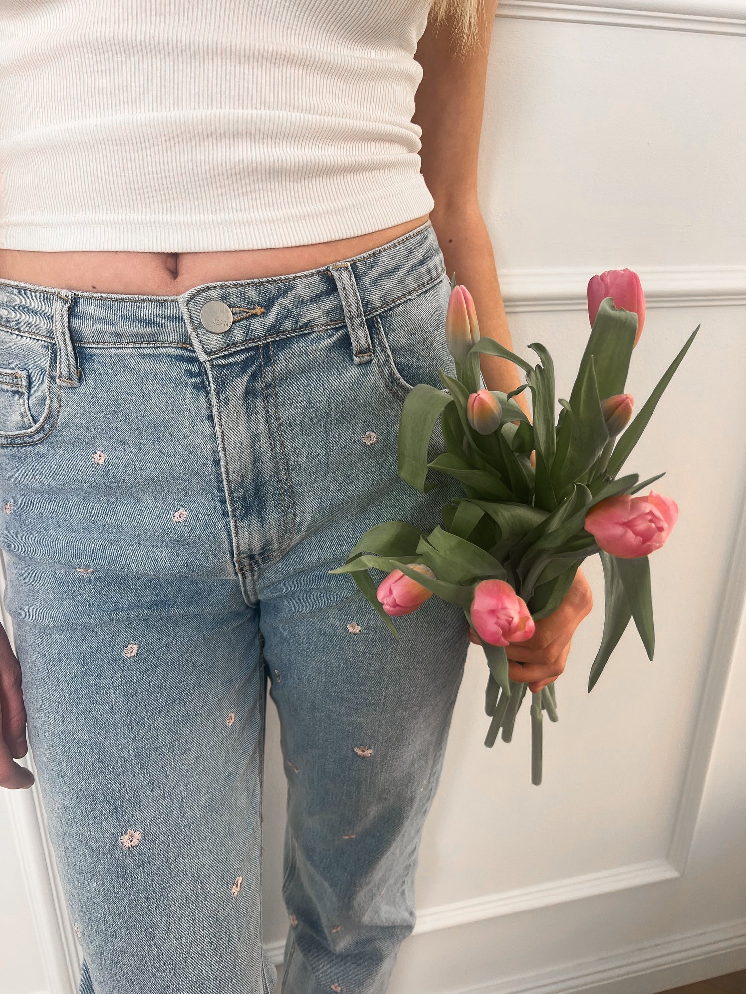 Frau hält pinke Blumen in der Hand und hält sie vor eine Jeans-Hose, die ebenfalls mit pinken Blumen verziert ist.
