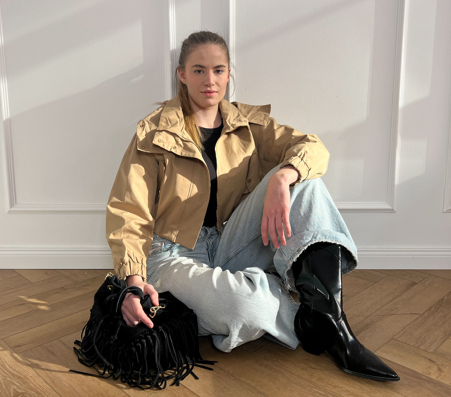 Person mit beiger Jacke und schwarzen Cowboyboots sind auf dem Boden und hat eine Tasche mit Fransen in der Hand