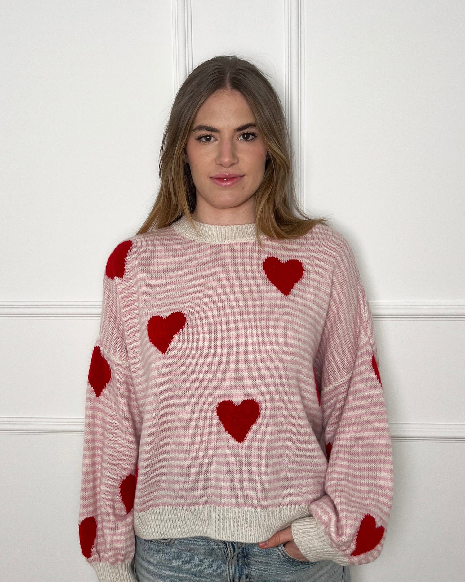 Blone Frau vor weißer Wand mit einem pink weiß gestreiften Pullover mit roten Herzen.