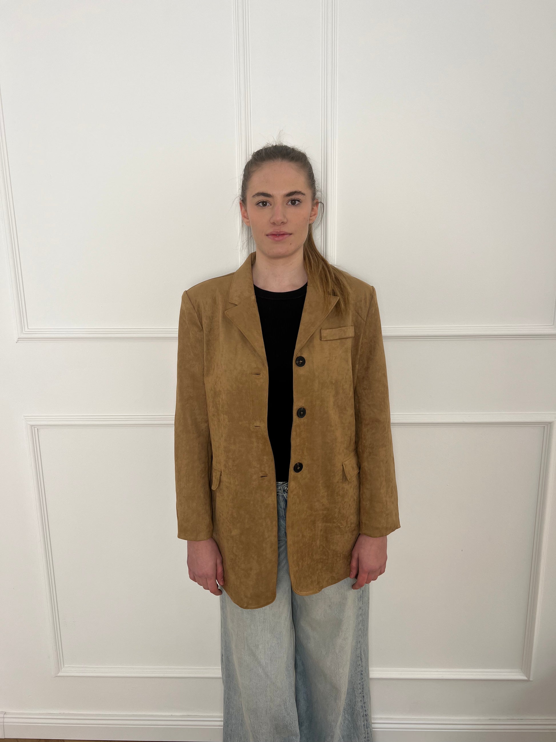 Brauner Blazer mit schwarzen Knöpfen getragen von einer Frau mit Zopf vor weißer Wand.