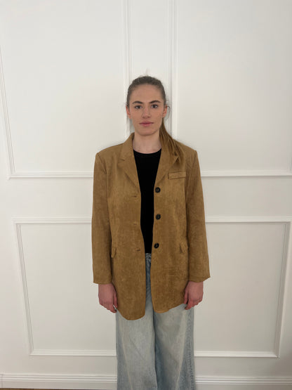 Brauner Blazer mit schwarzen Knöpfen getragen von einer Frau mit Zopf vor weißer Wand.