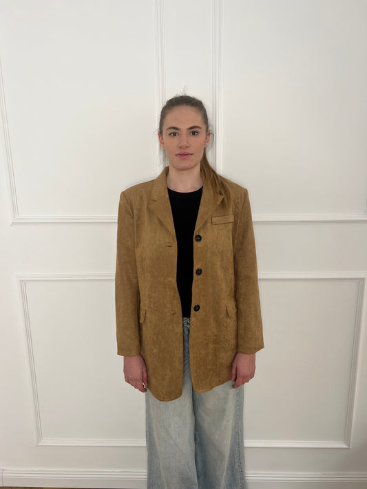 Brauner Blazer mit schwarzen Knöpfen getragen von einer Frau mit Zopf vor weißer Wand.