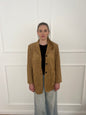 Brauner Blazer mit schwarzen Knöpfen getragen von einer Frau mit Zopf vor weißer Wand.