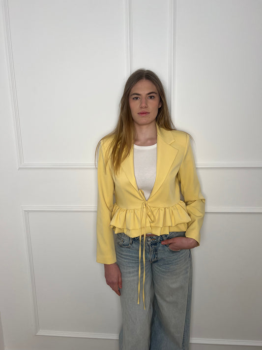 Blonde Frau vor weißer Wand mit einem gelben Blazer und Jeans
