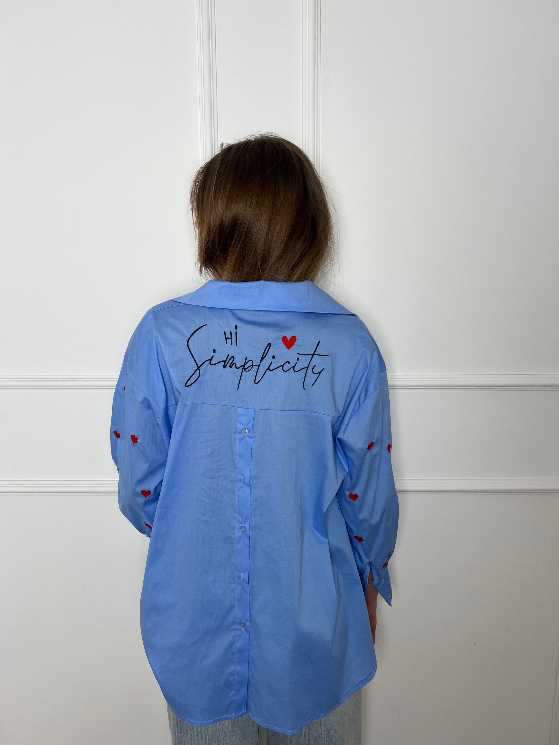 Frau steht vor einer weißen Wand von hinten und trägt eine blaue Bluse mit roten Herzen.