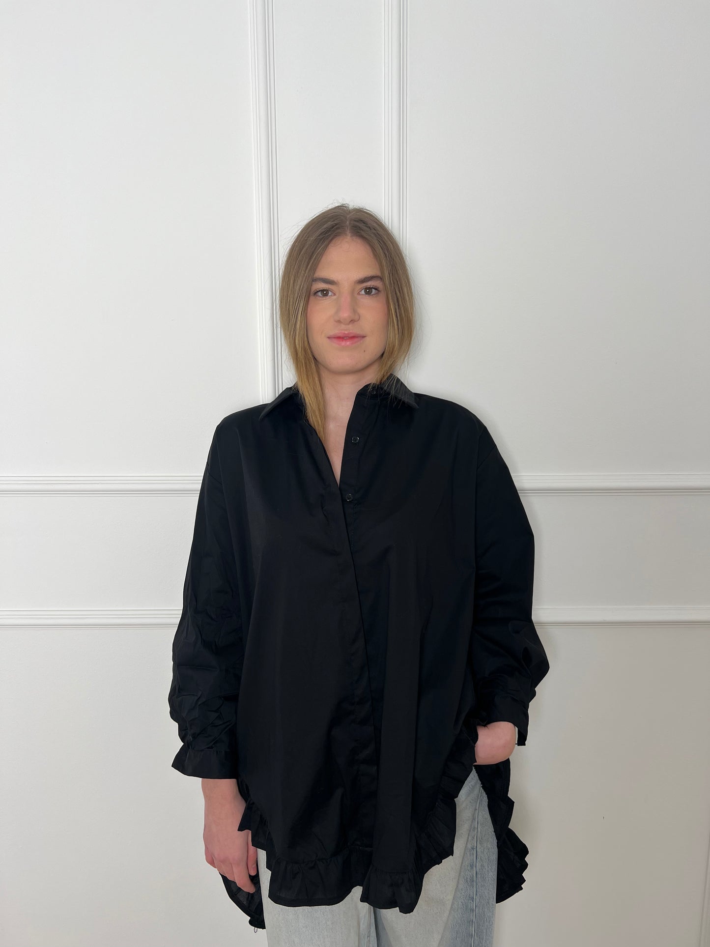 Blonde Frau vor weißer Wand trägt eine lange schwarze Bluse