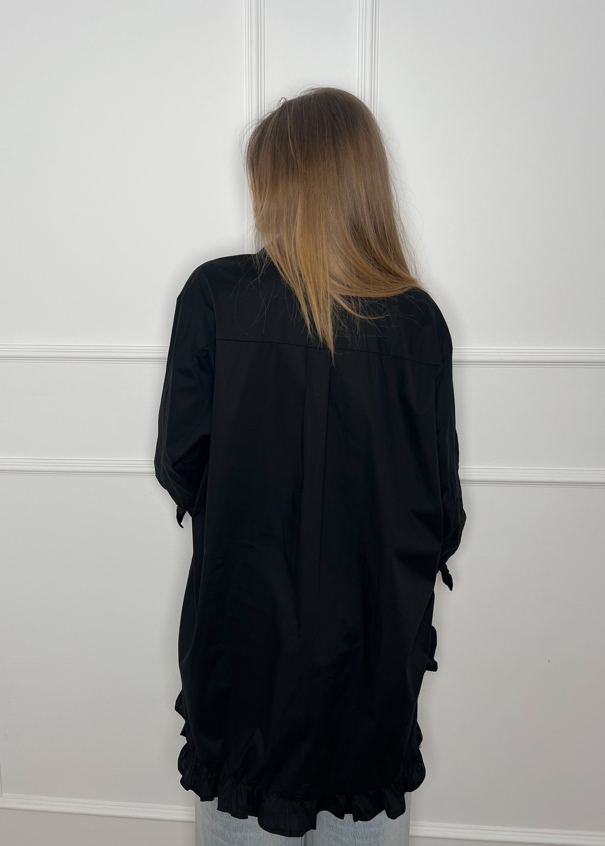 Blonde Frau vor weißer Wand trägt eine lange schwarze Bluse