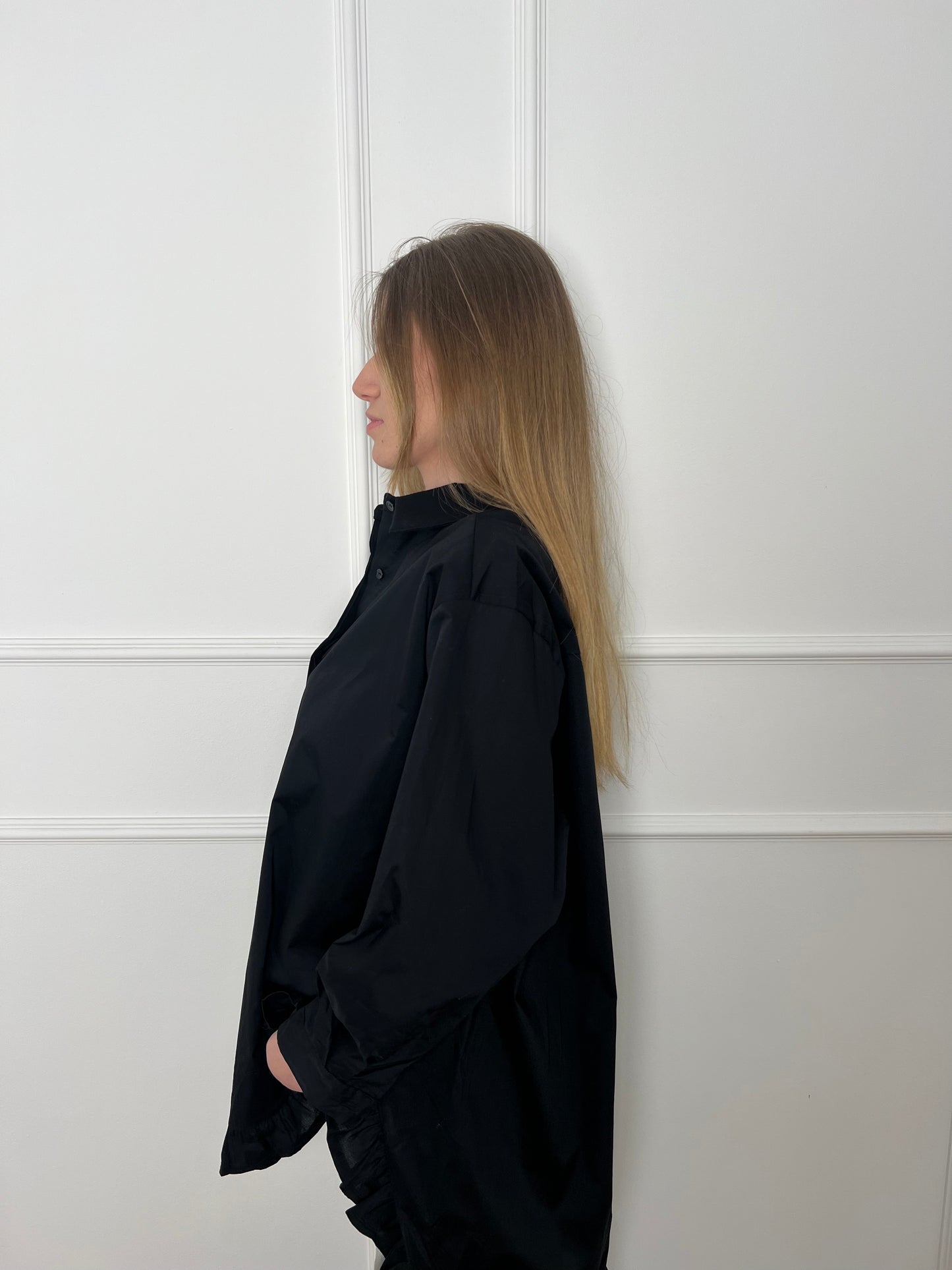 Blonde Frau vor weißer Wand trägt eine lange schwarze Bluse