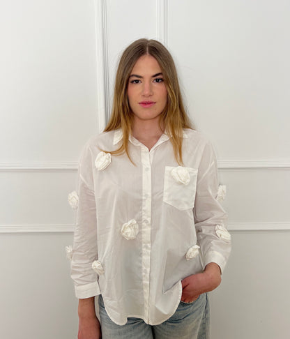 Blonde Frau vor weißer Wand mit einer weißen Bluse, die mit 3D Blumen besetzt ist. 