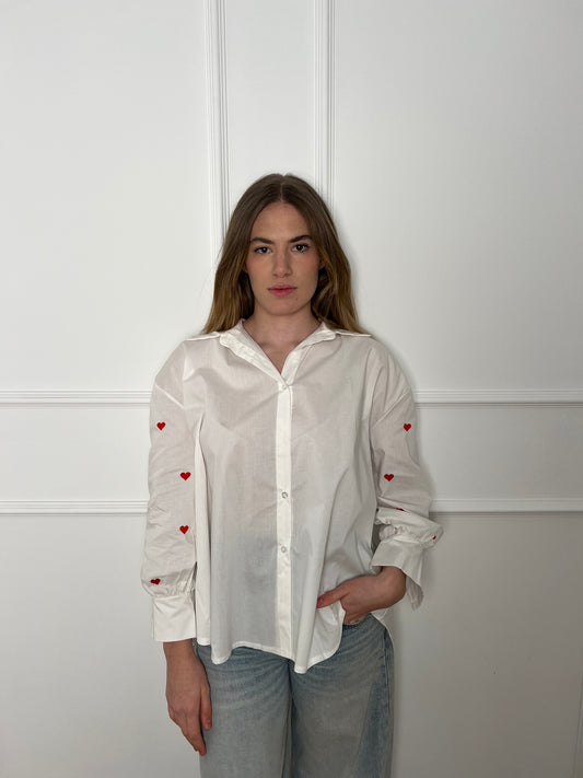 Blonde Frau vor weißer Wand trägt eine weiße Bluse, die an den Armen rote Herzen hat.