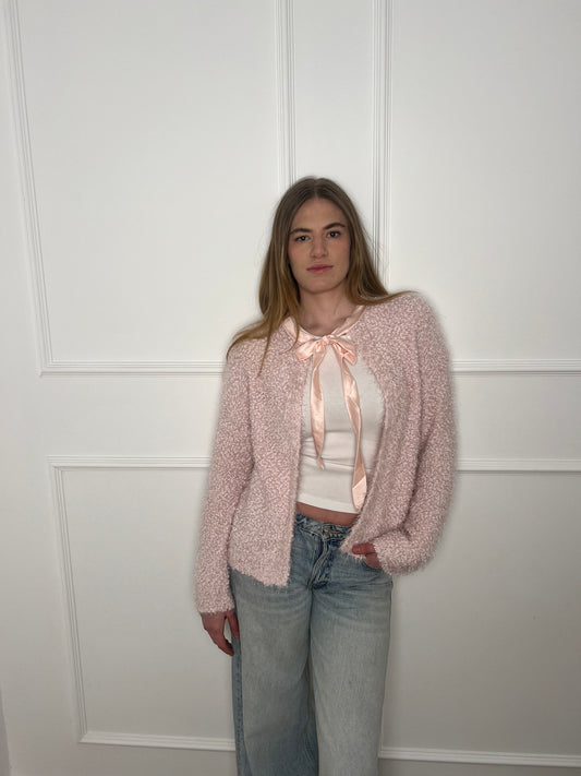 Blonde Frau vor weißer Wand mit rosanem Cardigan und Schleife