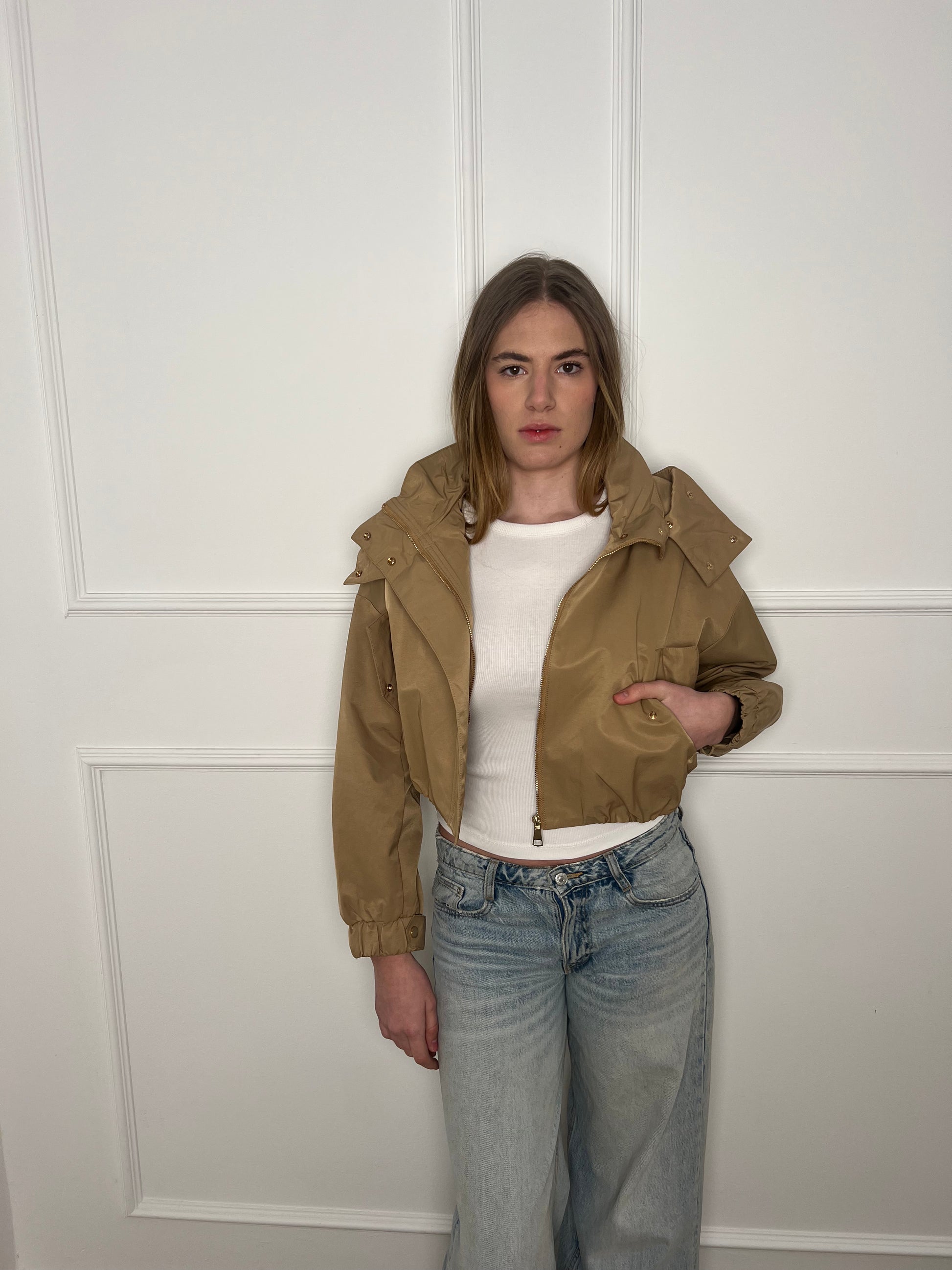 Frau vor weißer Wand trägt eine braune cropped Jacke und eine Jeans.
