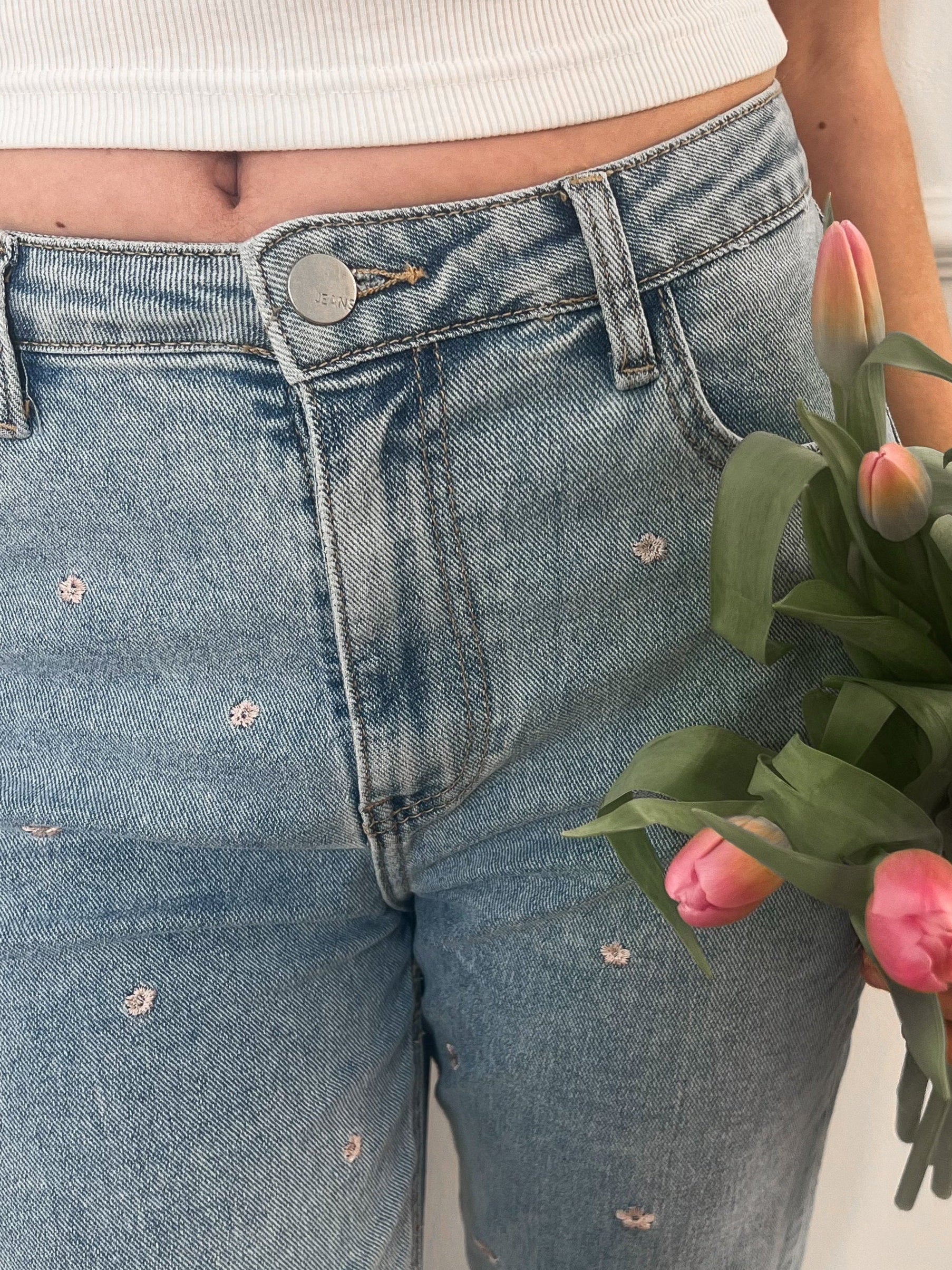 Blonde Frau trägt eine Jeans mit rosanen Blumen und hält einen Strauß pinke Tulpen in der Hand.