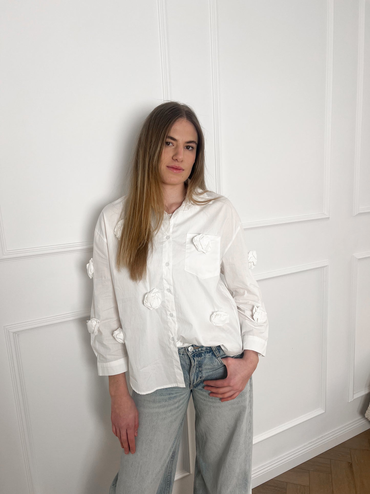 Blonde Frau posiert vor einer weißen Wand mit einer Jeans und einer weißen Bluse.