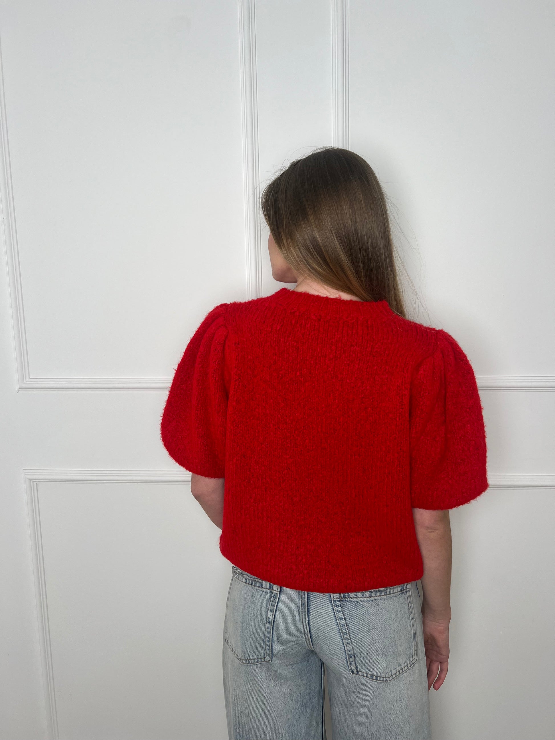 Frau von hinten vor weißer Wand trägt einen roten Pullover mit kurzen Armen.