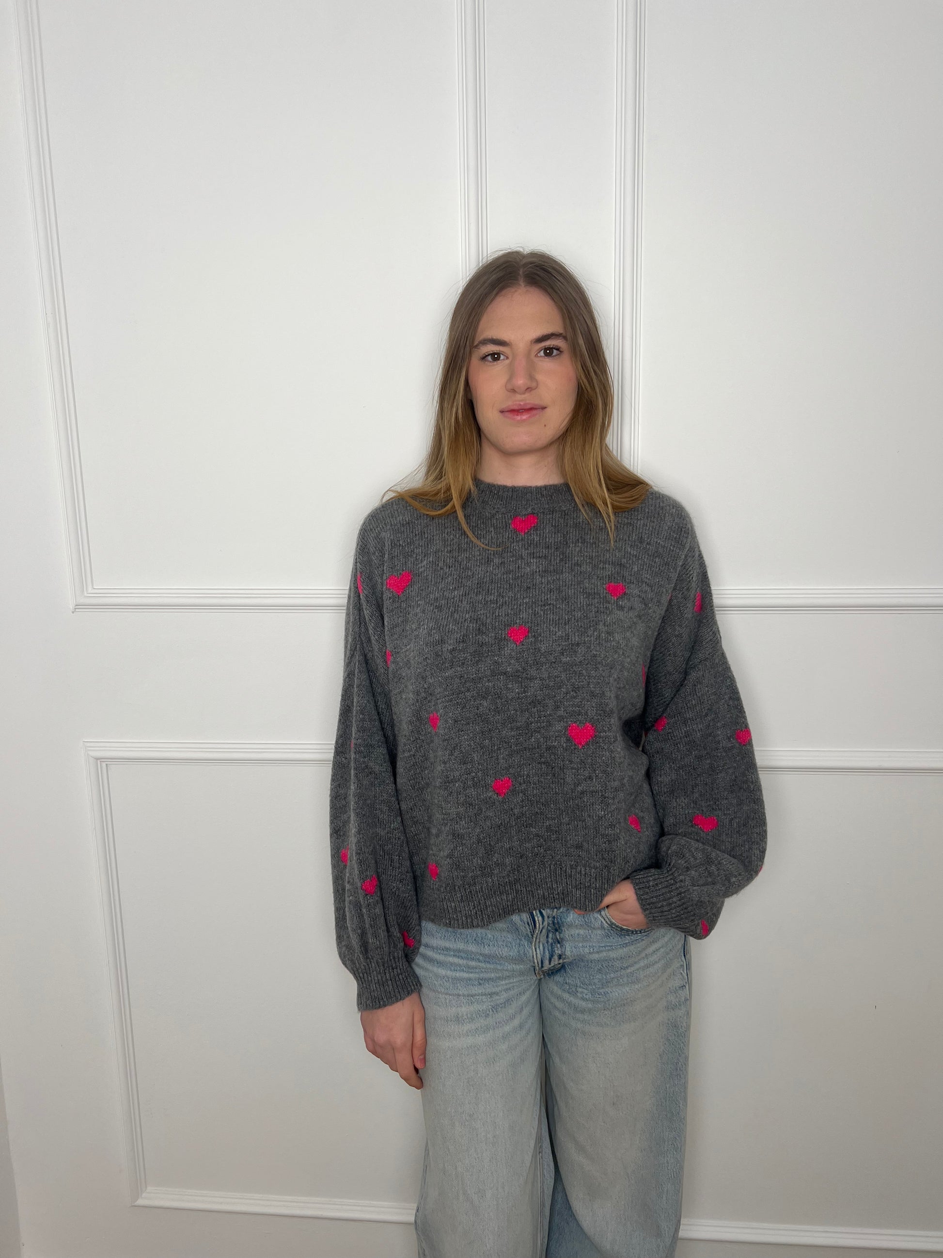 Frau vor weißer Wand trägt einen grauen Pullover mit pinken Herzen