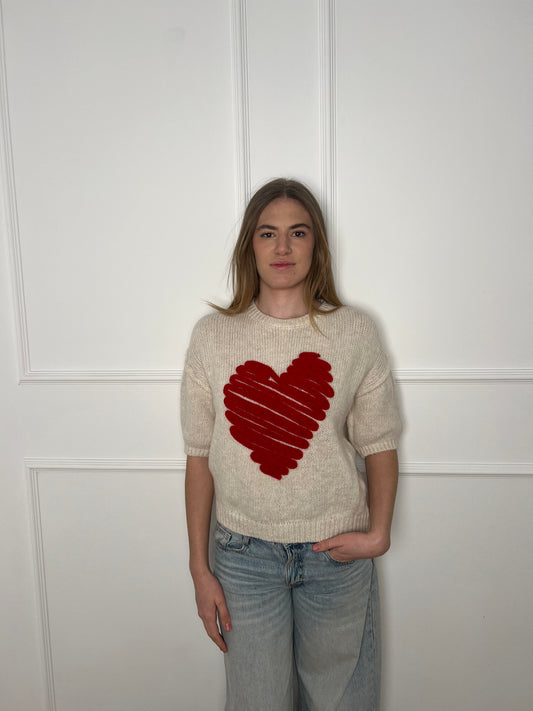 Frau vor weißer Wand trägt einen weissen Kurzarm-Pullover mit einem großen roten Herzen.
