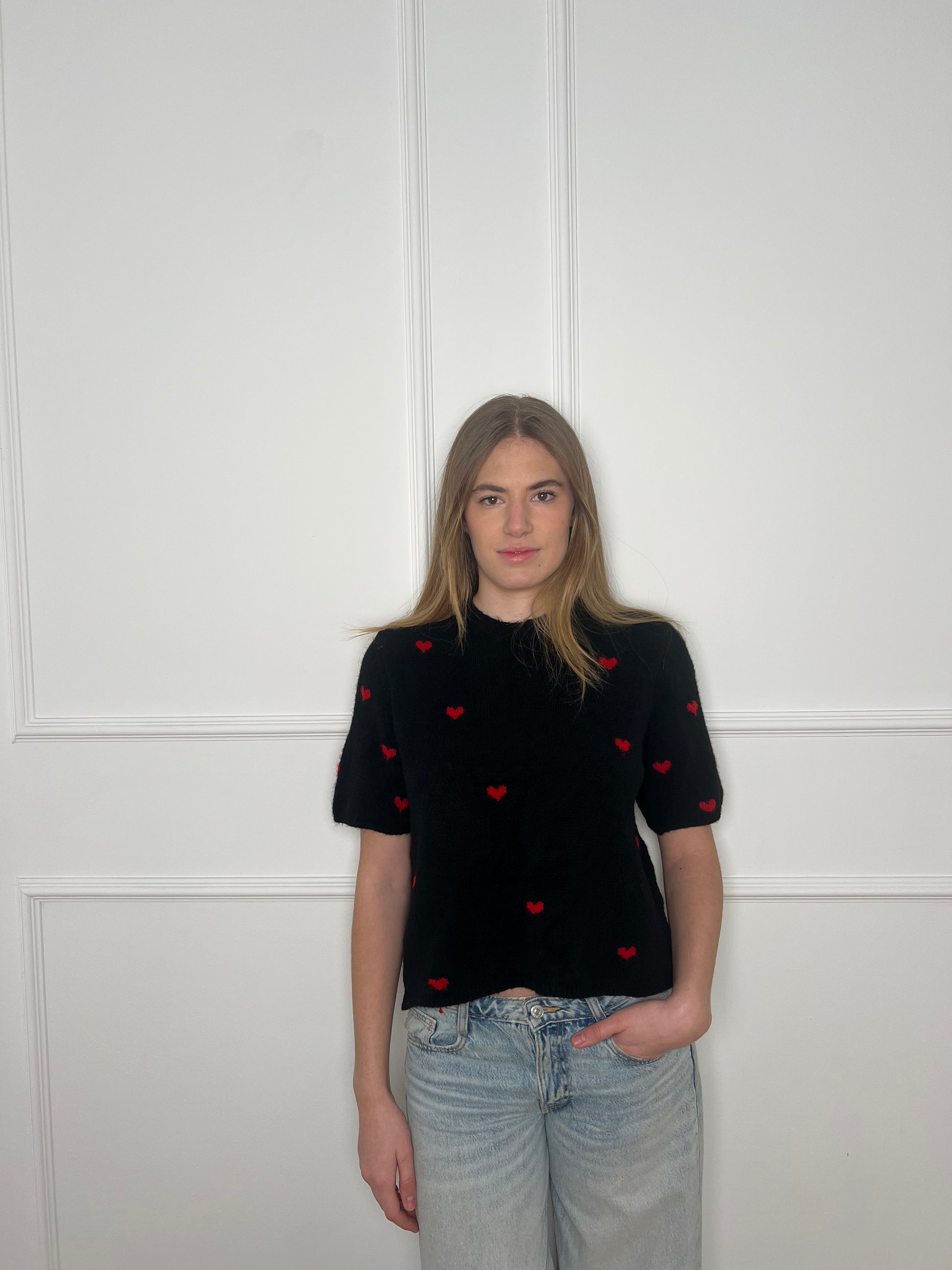 Frau vor weißer Wand trägt einen schwarzen Pullover mit kurzen Armen und roten Herzen.