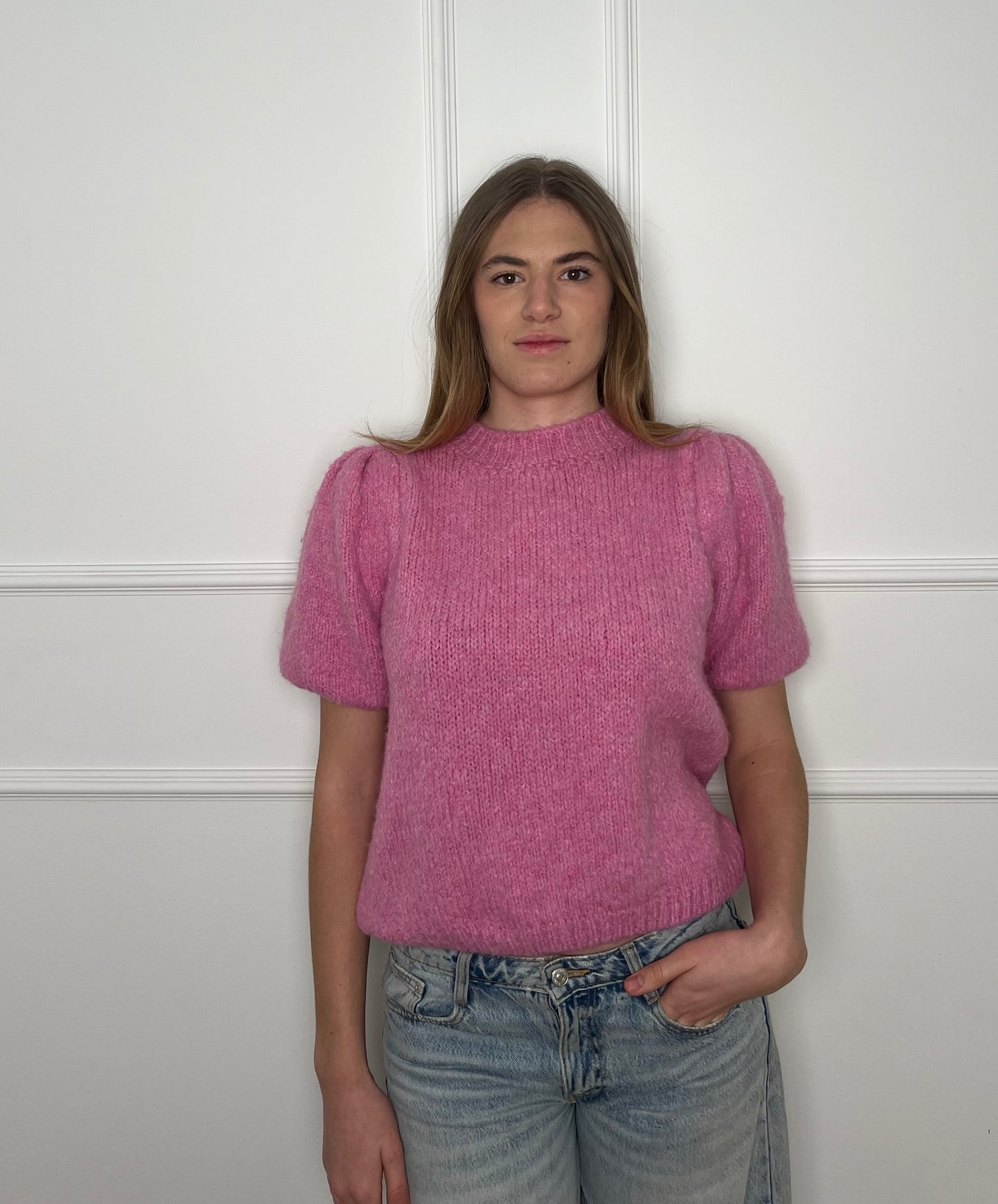 Frau vor weißer Wand trägt einen pinken Pullover mit kurzen Armen.