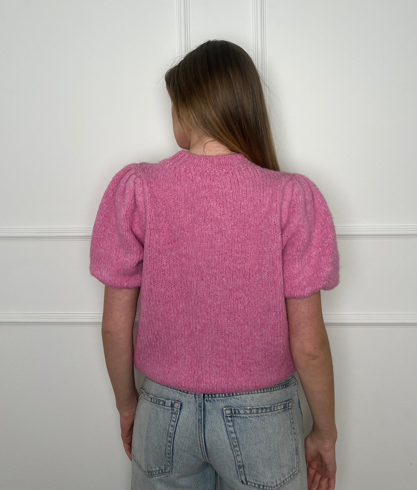Frau von hinten vor weißer Wand trägt einen pinken Pullover mit kurzen Armen.