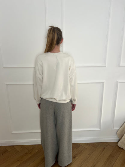 Frau vor weißer Wand trägt ein cremefarbenes Sweatshirt und eine graue Jogginghose