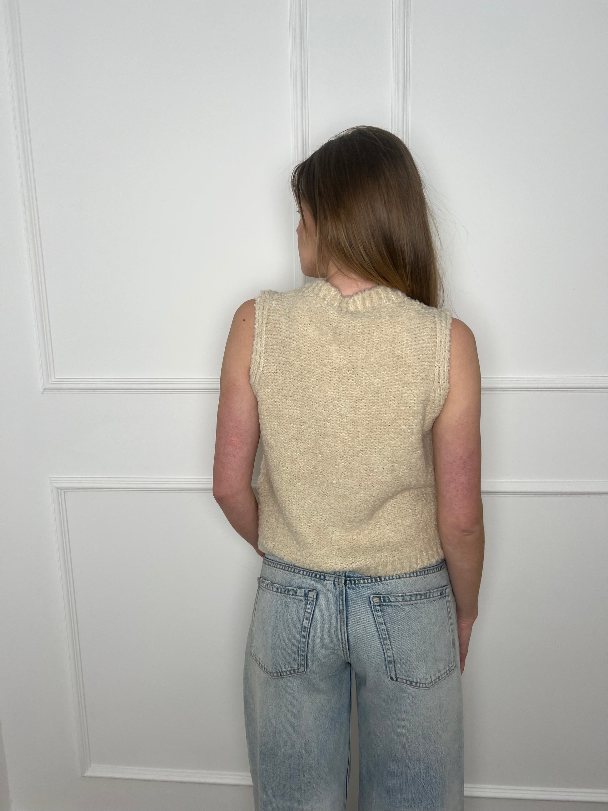 Frau vor weißer Wand trägt eine Weste in beige und ist von hinten zu sehen.