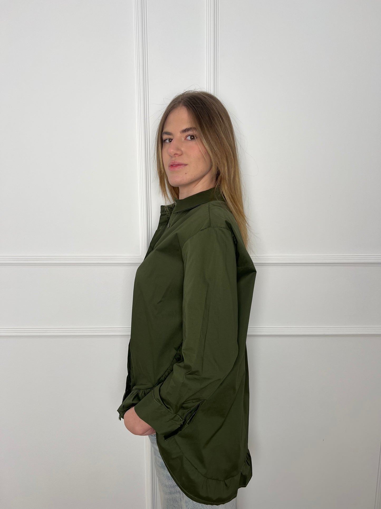 Blonde Frau steht seitlich vor weißer Wand und trägt eine olivgrüne Bluse