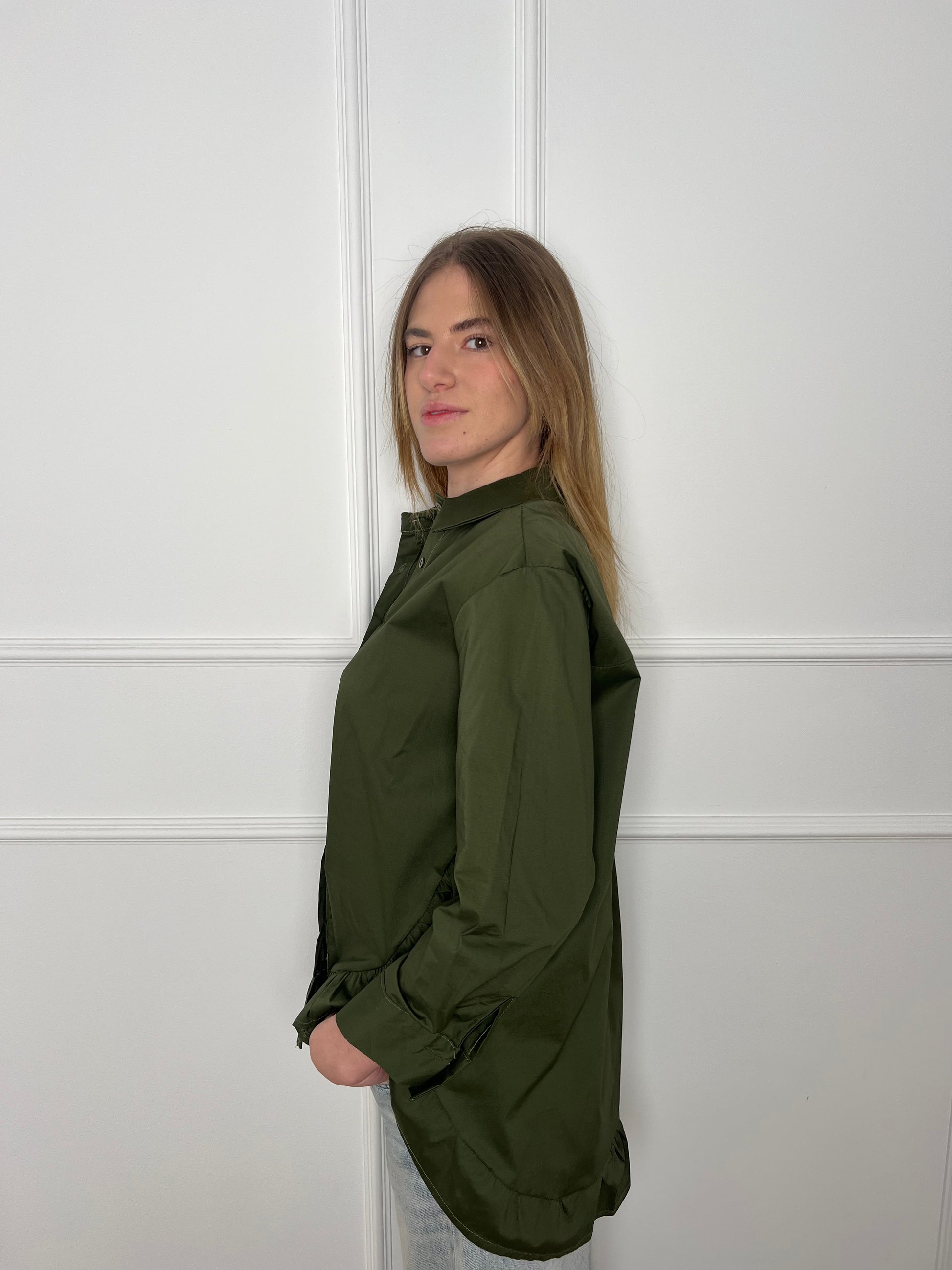 Blonde Frau steht seitlich vor weißer Wand und trägt eine olivgrüne Bluse