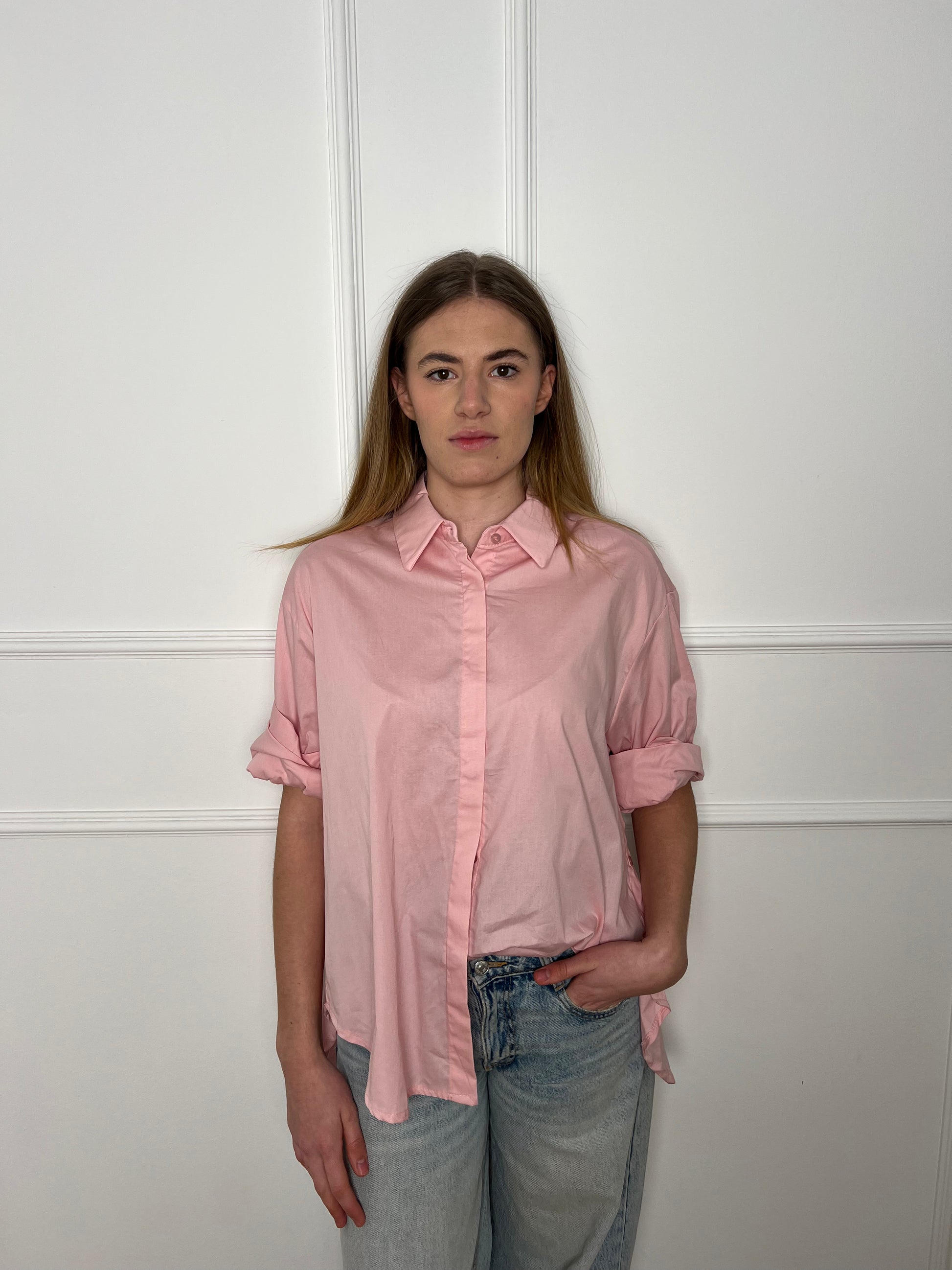 Blonde Frau vor weißer Wand trägt eine rosane Bluse, die in die Jeans gesteckt ist.