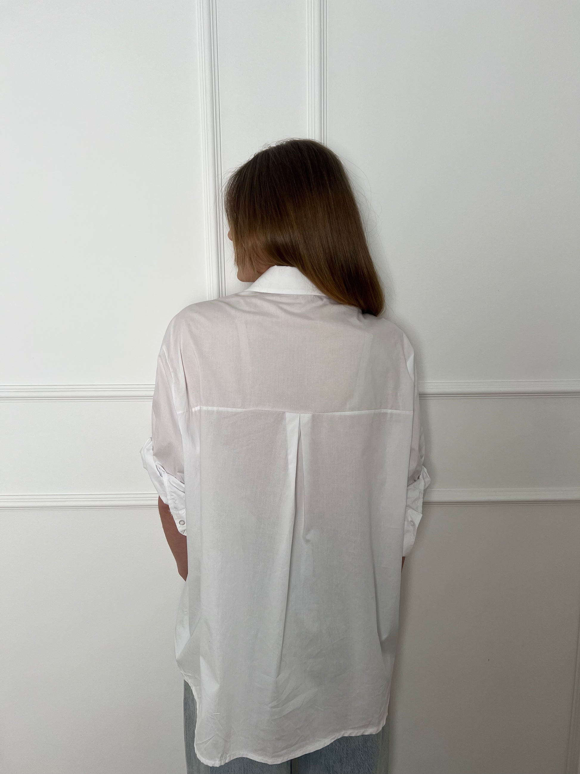 Blonde Frau vor weißer Wand trägt eine weiße Bluse und dreht den Rücken zu