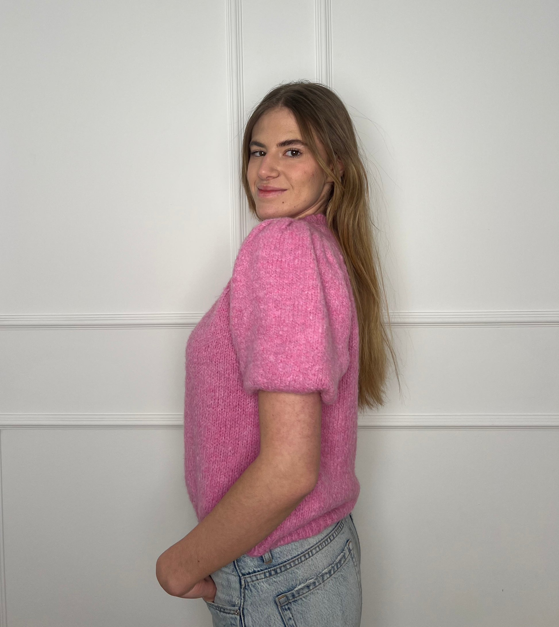 Frau von der Seite vor weißer Wand trägt einen pinken Pullover mit kurzen Armen.