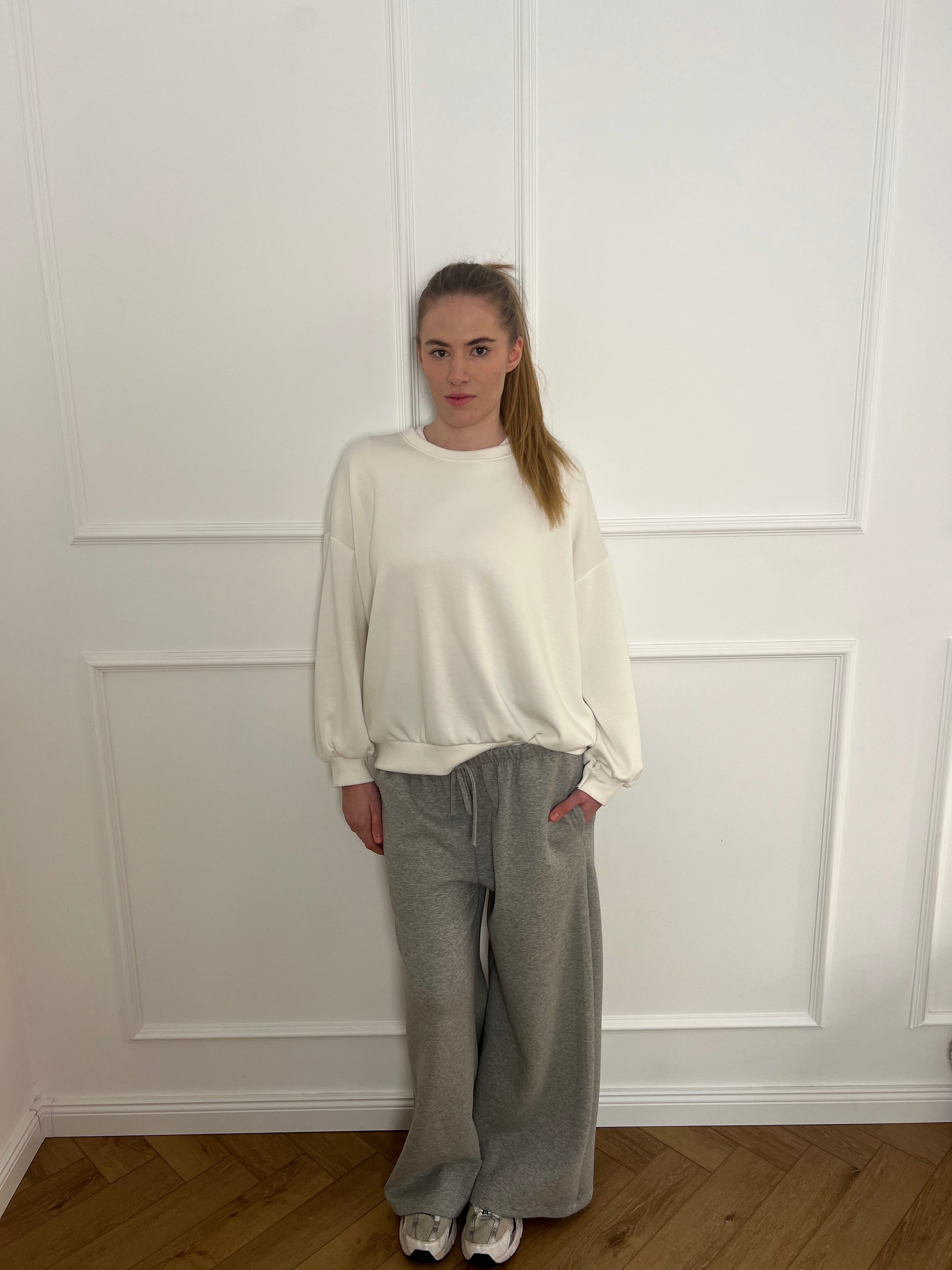 Frau vor weißer Wand trägt ein weißes Sweatshirt und eine graue Jogginghose